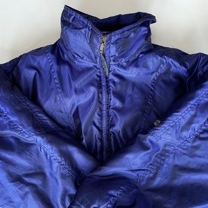 Op de afbeelding: Een felblauwe jas met een hoge kraag en een ritssluiting over de hele lengte. De jas heeft een gewatteerd ontwerp en een klein logo op de linkermouw. Gemaakt van een glanzend, lichtgewicht materiaal.