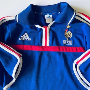 May include: A blue Adidas polo shirt featuring a white collar with blue trim, a red horizontal stripe across the chest, and white stripes along the sleeves. The shirt displays an embroidered gold rooster emblem above the F.F.F. logo and a star.