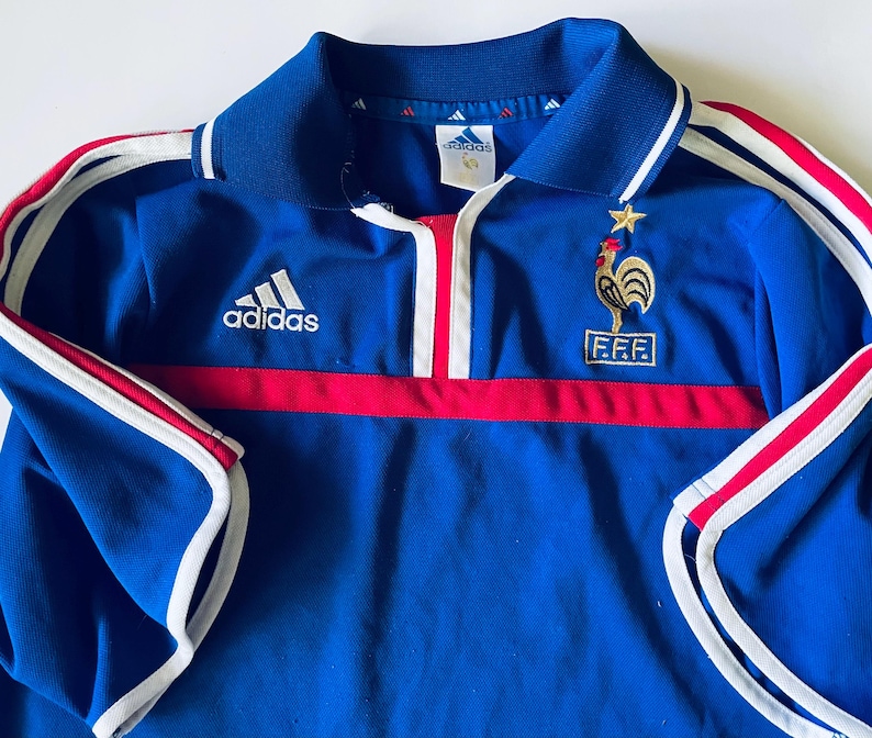 May include: A blue Adidas polo shirt featuring a white collar with blue trim, a red horizontal stripe across the chest, and white stripes along the sleeves. The shirt displays an embroidered gold rooster emblem above the F.F.F. logo and a star.