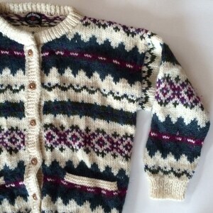 Op de afbeelding: Crèmekleurig vest met een geometrisch patroon in teal, paars en wit. De trui heeft een knoopsluiting, twee voorzakken en geribde manchetten. De merknaam "Lost Horizons" is zichtbaar op het label.