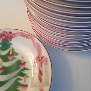 Op de afbeelding: Een stapel witte keramische borden met roze en groene kerstontwerpen. De borden hebben een kerstboom met ornamenten, een cadeaudoos en een lint.