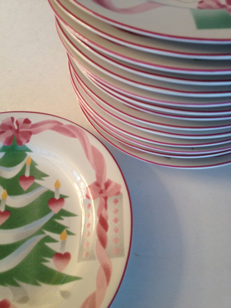 Op de afbeelding: Een stapel witte keramische borden met roze en groene kerstontwerpen. De borden hebben een kerstboom met ornamenten, een cadeaudoos en een lint.