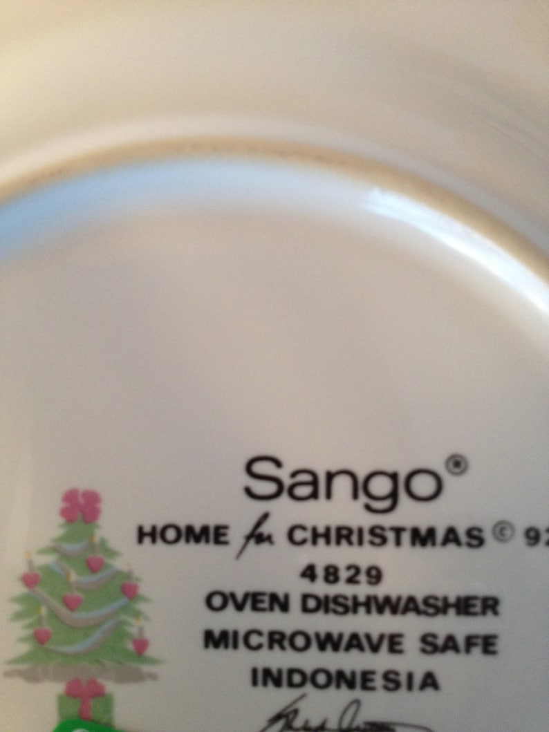 Op de afbeelding: Een wit keramiek bord met een kerstboomontwerp en de tekst "Sango&reg; HOME for CHRISTMAS&reg; 92 4829 OVEN DISHWASHER MICROWAVE SAFE INDONESIA".