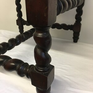 May include: Close-up of a dark brown wooden table leg with a twisted design. The leg is square at the top and bottom, with a rounded, twisted section in the middle.