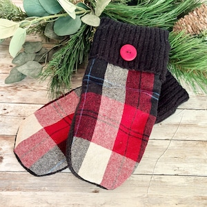 May include: A pair of handmade mittens with a red, black, and white plaid pattern. The cuffs are dark gray knit with a red button detail. The mittens are displayed on a wooden surface with greenery in the background.