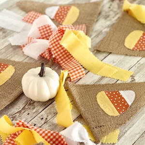 May include: A festive fall banner with burlap pennants, each adorned with a candy corn design. The banner is accented with yellow, white, and orange gingham ribbons, and a small white pumpkin. Perfect for autumn decor.