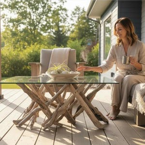 May include: A coffee table featuring a clear rectangular glass top supported by a base constructed from numerous light-coloured, intertwined driftwood branches. The table is positioned on a wooden deck, showcasing a natural, organic aesthetic.