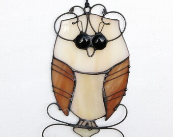 May include: A stained glass owl with brown and white wings, black eyes, and a wire frame. The owl is hanging from a black cord.