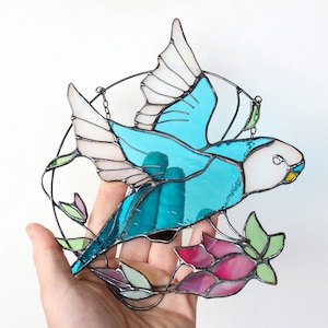 May include: A stained glass suncatcher featuring a blue bird with white and pink wings, surrounded by green and pink floral details.