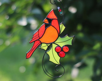 May include: A stained glass cardinal perched on a holly branch with red berries. The cardinal is red, orange, and black. The holly leaves are green and yellow.
