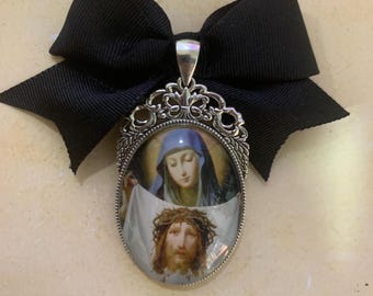 Op de afbeelding: Een religieuze hanger met een ovale afbeelding van Jezus en Maria, gezet in een sierlijk zilverkleurig frame. De hanger is versierd met een grote zwarte strik aan de bovenkant. De afbeelding toont Jezus met een doornenkroon en Maria in een blauwe sluier.
