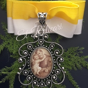 Puede incluir: Un colgante de plata con un diseño filigrana detallado presenta una imagen de camafeo de San Antonio de Padua sosteniendo a un bebé Jesús. El colgante está unido a una cinta amarilla y blanca.