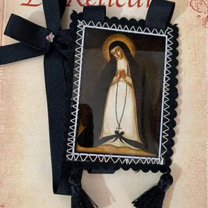 Può includere: Un oggetto devozionale religioso con l'immagine di un santo, incorniciato con un bordo smerlato nero e cuciture bianche. Ha un nastro nero con un fiocco e un piccolo gioiello, più due nappe nere.