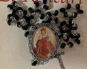 May include: A rosary with black beads and a silver-toned crucifix. The centerpiece features an oval image of a religious figure in a red and gold gown. The words "El Relicari" are visible in the background.
