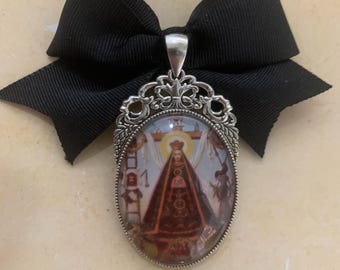 Op de afbeelding: Een ovale hanger met een religieuze afbeelding, omlijst in sierlijk zilvermetaal, is bevestigd aan een zwarte strik. De hanger toont een centrale figuur in een donkere jurk, omringd door andere figuren. De achtergrond is lichtbruin.
