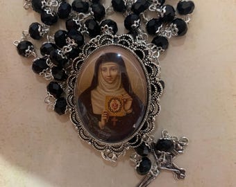 May include: A rosary with black beads and a silver-toned crucifix. The centerpiece features an oval image of a saint within an ornate frame. The rosary is likely used for prayer and religious devotion.