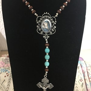 May include: A brown wooden rosary necklace with turquoise beads and a silver pendant featuring a depiction of the Virgin Mary. The pendant is attached to a silver crucifix.
