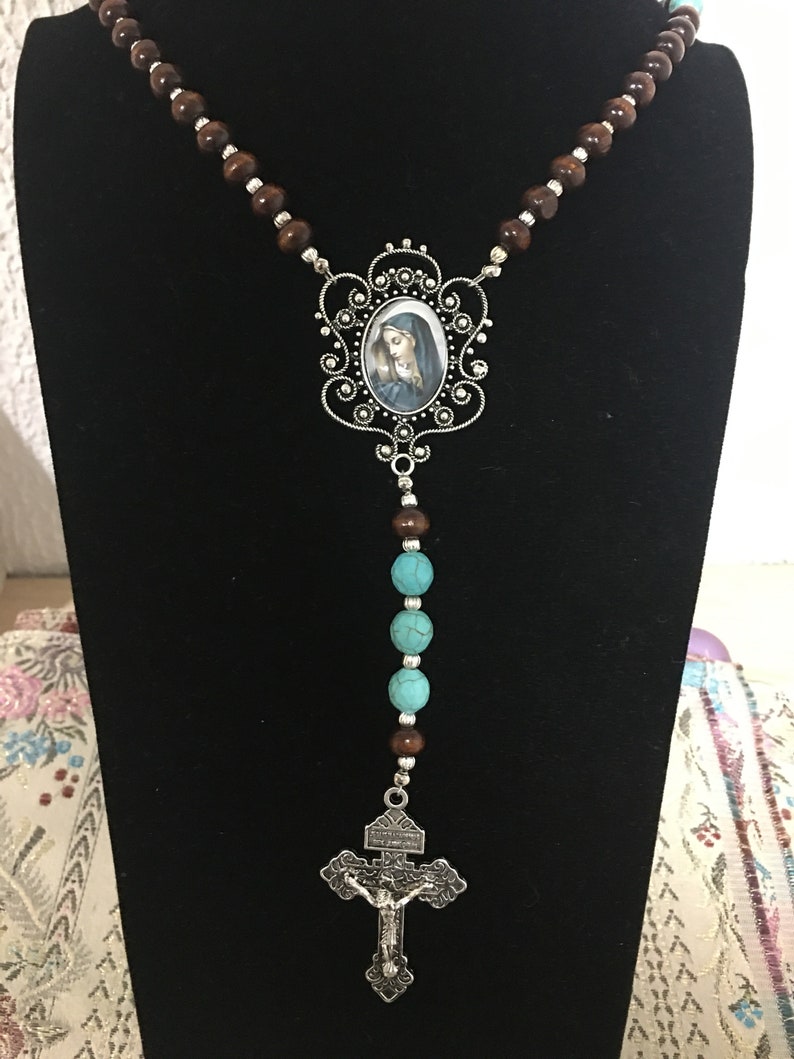 May include: A brown wooden rosary necklace with turquoise beads and a silver pendant featuring a depiction of the Virgin Mary. The pendant is attached to a silver crucifix.