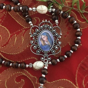 May include: A brown wooden rosary with white beads and a silver pendant featuring a blue and white image of the Virgin Mary.