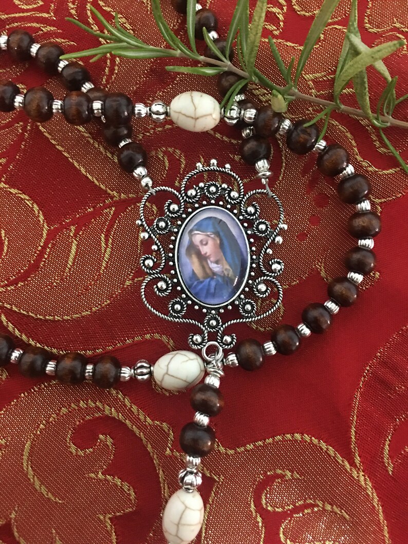 May include: A brown wooden rosary with white beads and a silver pendant featuring a blue and white image of the Virgin Mary.