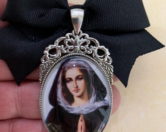 Op de afbeelding: Een ovale hanger met een religieuze afbeelding van een vrouw, gezet in een zilverkleurig montuur, bevestigd aan een zwarte strik. De hanger heeft een religieuze afbeelding. Het item is religieus gethematiseerd.