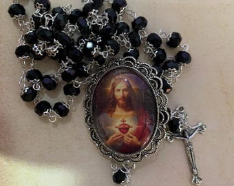 May include: A rosary with black faceted beads and a silver-toned crucifix. The centerpiece features an oval pendant with an image of Jesus. The text "El Relicari" is visible in the background.