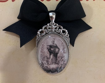 Op de afbeelding: Een ovale hanger met een religieuze afbeelding, omlijst in sierlijk zilverkleurig metaal, is bevestigd aan een zwarte strik. De hanger heeft een zwart-wit illustratie van een heilige. De achtergrond heeft de rode woorden "El Relicario".