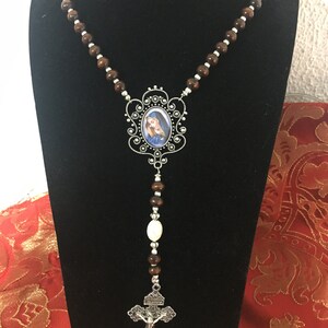 May include: A brown wooden rosary with a silver crucifix and a silver oval pendant featuring a picture of the Virgin Mary. The rosary is displayed on a black stand.
