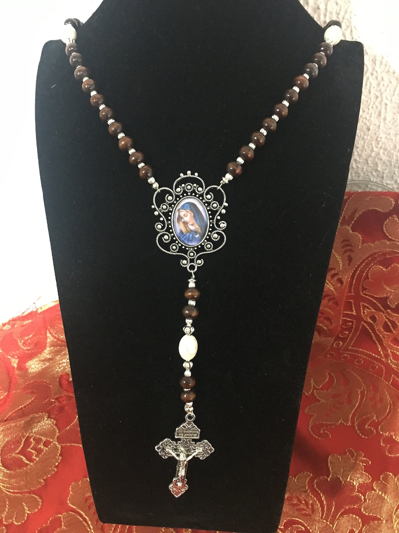 May include: A brown wooden rosary with a silver crucifix and a silver oval pendant featuring a picture of the Virgin Mary. The rosary is displayed on a black stand.