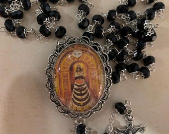 May include: A rosary with black faceted beads and a silver-toned crucifix. The centerpiece features an oval pendant with a religious image. The rosary is displayed on a surface with the text "El Relicari".