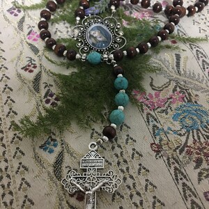 May include: A brown wooden rosary with turquoise beads and a silver crucifix. The crucifix has the words "JESUS MADE ME HIS,  REY DE GLORIA" on it. The rosary also has a silver pendant with a picture of the Virgin Mary.