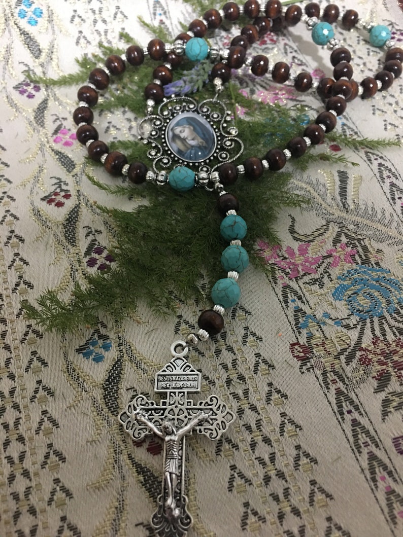 May include: A brown wooden rosary with turquoise beads and a silver crucifix. The crucifix has the words "JESUS MADE ME HIS,  REY DE GLORIA" on it. The rosary also has a silver pendant with a picture of the Virgin Mary.