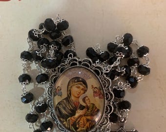 May include: A rosary with black beads and a silver-toned crucifix. The centerpiece features an oval image of Mary holding baby Jesus. The text "El Relicari" is visible in the background.