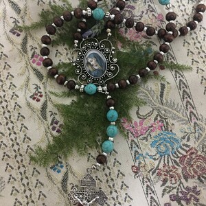 May include: A brown wooden rosary with turquoise beads and a silver pendant featuring a picture of the Virgin Mary. The pendant is attached to a silver cross with a tree design.