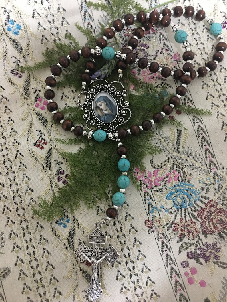 May include: A brown wooden rosary with turquoise beads and a silver pendant featuring a picture of the Virgin Mary. The pendant is attached to a silver cross with a tree design.