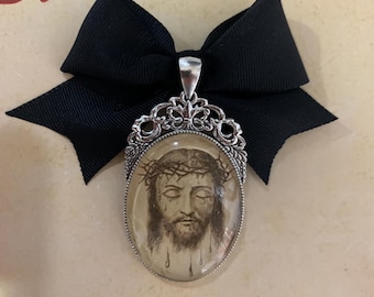 Op de afbeelding: Een ovale hanger met een afbeelding van Jezus met een doornenkroon, gezet in een zilverkleurig montuur. Bevestigd aan een zwarte strik. De achtergrond toont de tekst "El Relicario".