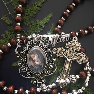 May include: A brown wooden rosary with silver beads and a silver pendant featuring a picture of the Virgin Mary and a silver crucifix with the text "JESUS NAZARENO REY DE REYES" on the crossbar.