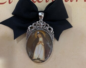 Op de afbeelding: Een ovale hanger met een religieuze afbeelding, omlijst in sierlijk zilverkleurig metaal, hangt aan een zwarte strik. De afbeelding toont een vrouw in een witte jurk. De hanger is bevestigd aan een zilverkleurige beugel.