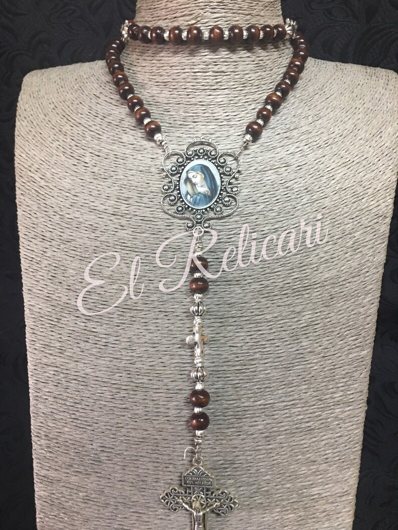 May include: A brown wooden rosary with a silver pendant featuring a depiction of the Virgin Mary. The pendant is surrounded by an ornate silver frame. The rosary also has a silver crucifix with the inscription "Ecce Homo Filius Dei Salvator" at the bottom.