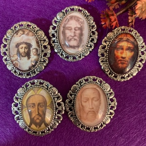 Könnte beinhalten: Fünf ovale religiöse Broschen mit silberfarbenen Filigranrahmen. Jede Brosche zeigt ein anderes Bild von Jesus Christus, mit unterschiedlichen Hauttönen und Gesichtsausdrücken. Der Hintergrund ist tiefviolett.
