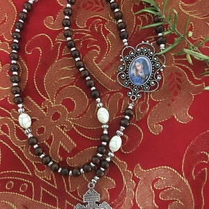 May include: A brown wooden rosary with white beads and a silver pendant featuring a picture of the Virgin Mary. The pendant is attached to a silver crucifix.