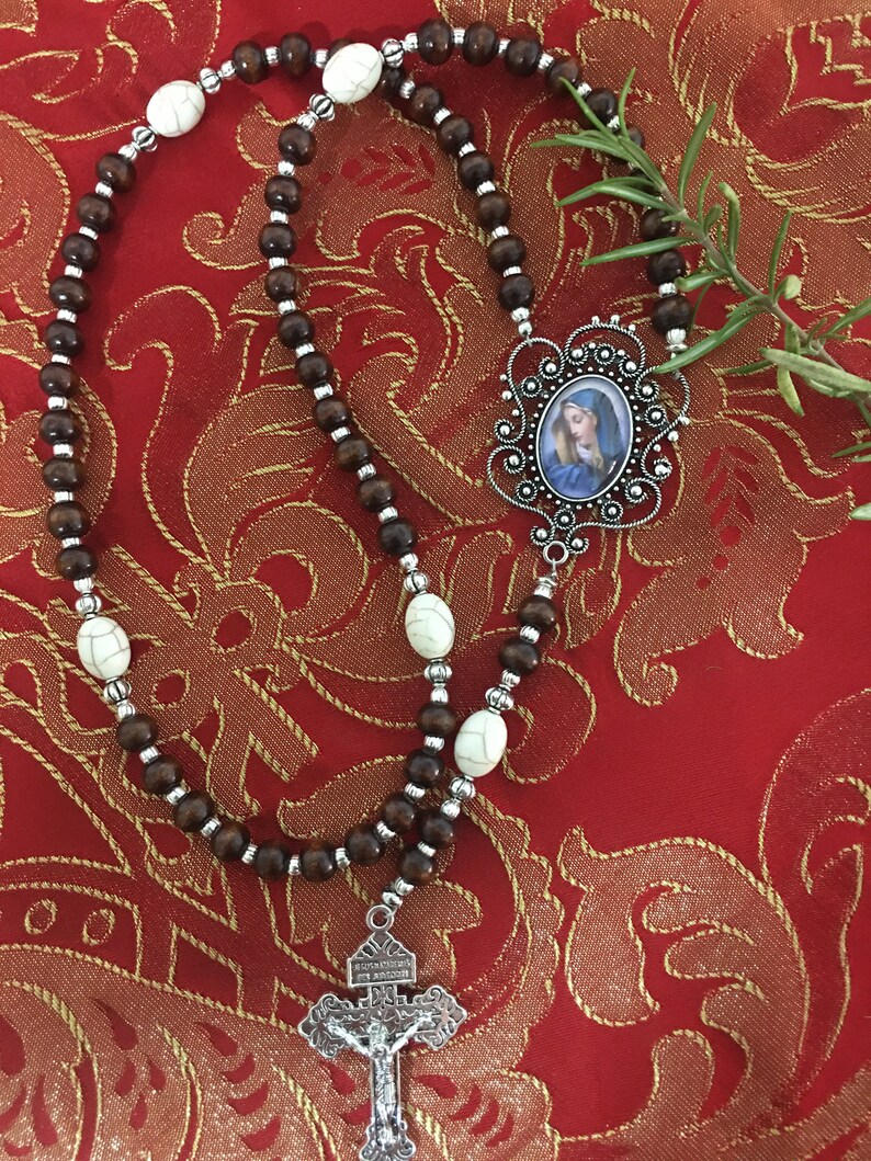 May include: A brown wooden rosary with white beads and a silver pendant featuring a picture of the Virgin Mary. The pendant is attached to a silver crucifix.