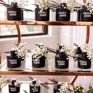 May include: Black cylindrical diffusers with the text "CHARLI DAWN" in white, arranged on a tiered gold and glass display. Each diffuser is adorned with a small bouquet of white flowers and black reeds.