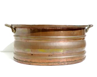 May include: An antique copper-coloured metal tub, oval in shape, with two small handles. The tub features horizontal bands around its exterior. The metal has an aged appearance, with variations in colour and patina.