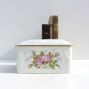 Pu&ograve; includere: Scatola in porcellana bianca con bordo dorato e un motivo floreale di rose rosa.