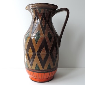 May include: A brown ceramic pitcher with a geometric pattern in shades of brown, black, and green. The pitcher has a wide mouth and a handle. The base of the pitcher is decorated with an orange band.