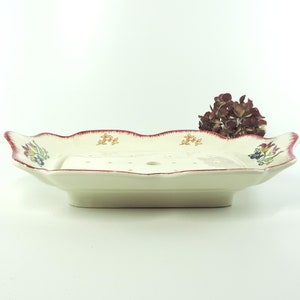 Puede incluir: Un jabonera de cerámica blanca con un borde rojo y un diseño floral. La jabonera tiene una base elevada y agujeros de drenaje.