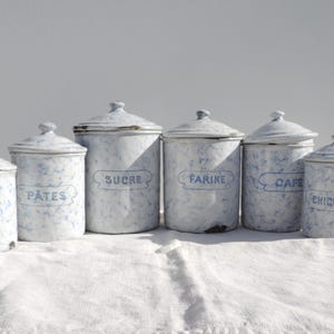 Puede incluir: Un conjunto de seis botes de almacenamiento de esmalte vintage en blanco con motas azules y tapas. Cada bote tiene una etiqueta azul con texto en francés: PÂTES, SUCRE, FARINE, CAFÉ y CHICORÉE. Los botes están colocados sobre un paño blanco.