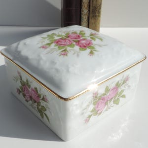 Pu&ograve; includere: Una scatola quadrata in porcellana bianca con un bordo dorato e un motivo floreale con rose rosa. La scatola ha una forma quadrata e un design in rilievo sul coperchio.