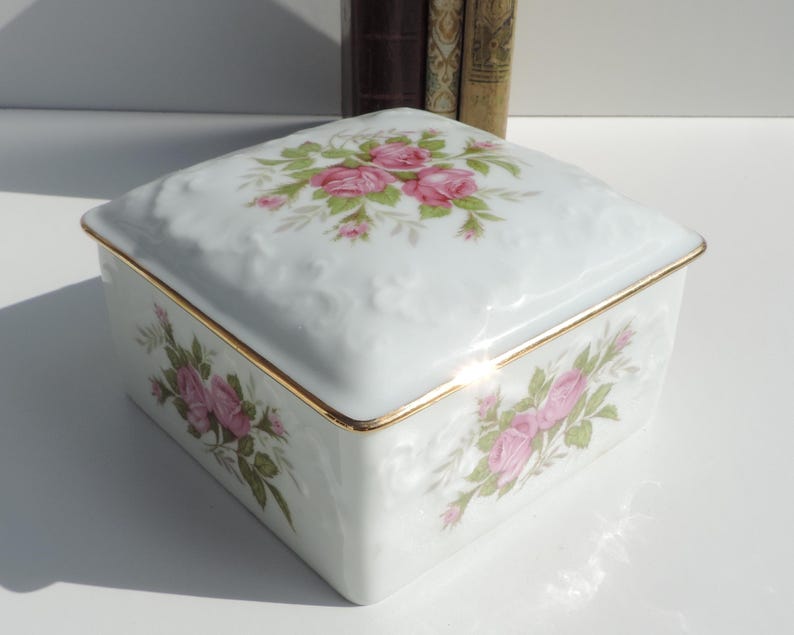 Pu&ograve; includere: Una scatola quadrata in porcellana bianca con un bordo dorato e un motivo floreale con rose rosa. La scatola ha una forma quadrata e un design in rilievo sul coperchio.